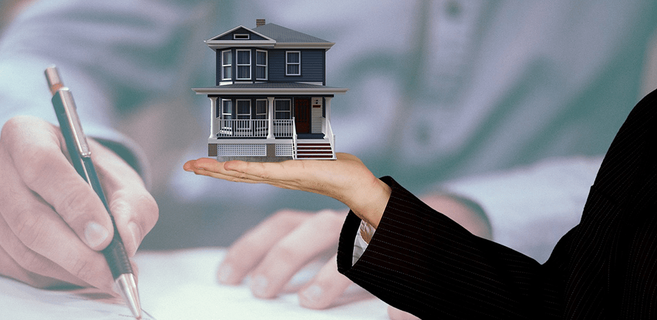 hand holding a model home with someone signing papers in the background